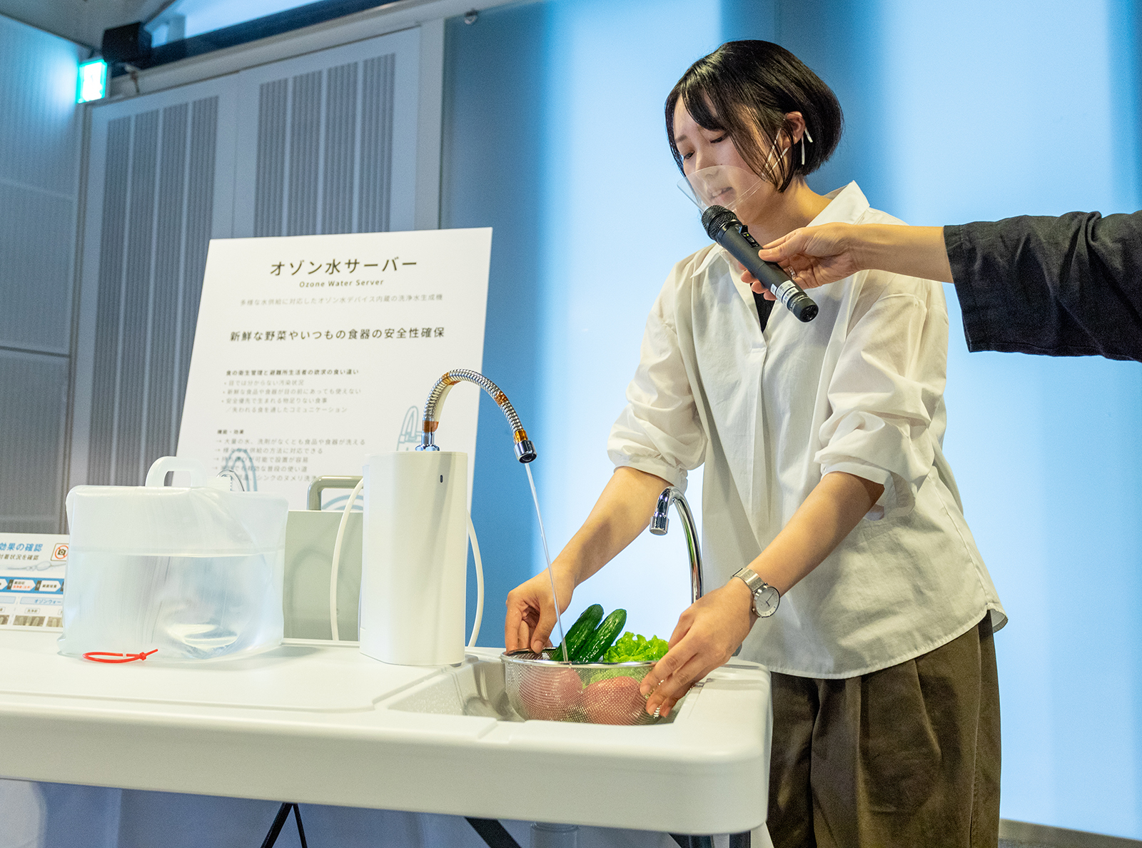 写真：「オゾン水サーバー」の使用シーンを実演する、学生の安藤 秋穂（あんどう あきほ）氏。